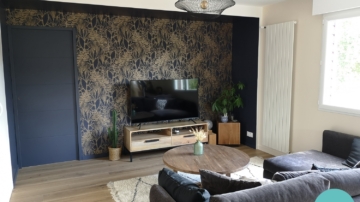 Salon avec papier peint bleu et or Casamance, peinture bleu nuit, meuble TV bois, canapé gris velours, table basse ronde bois, tapis berbère blanc et noir, suspension filaire noir métal