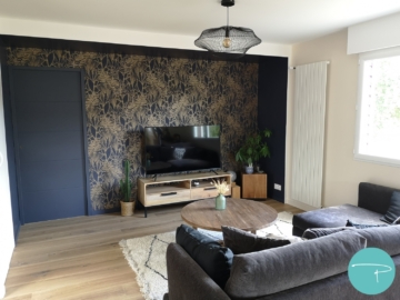 Salon avec papier peint bleu et or Casamance, peinture bleu nuit, meuble TV bois, canapé gris velours, table basse ronde bois, tapis berbère blanc et noir, suspension filaire noir métal