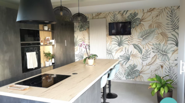 Rénovation d'une maison avec salon et salle à manger mur bleu vert, cuisine style industriel et papier peint panoramique feuillage, chambre beige et vert avec papier peint végétal, WC vert avec papier peint effet tasseaux bois, luminaires métal noir industriel grillage